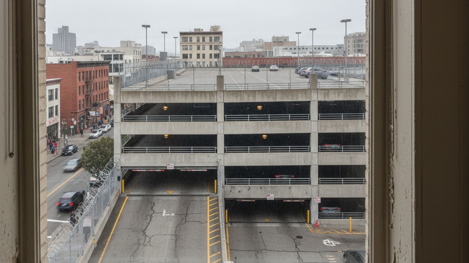A view down onto a city street from a few floors up, seen past the painted reveal of a tall office window, with the open decks of a multi storey parking structure on the far side of the road, flat grey daylight
